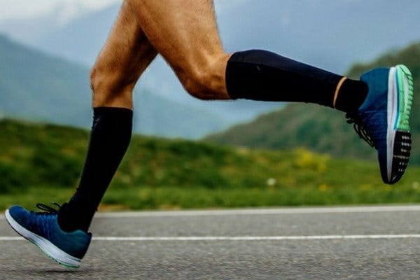 Ansicht zweier Beine eines Mannes, der in schnellem Tempo über eine asphaltierte Landstraße läuft. Er trägt Kompressionsstrümpfe und Laufschuhe. 