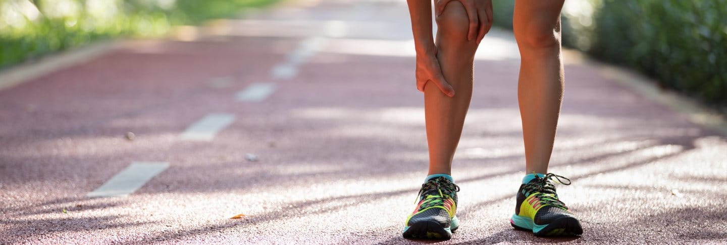Eine Frau im Sportoutfit steht vornüber gebeugt auf einer Landstraße und hält sich ihr schmerzendes Läuferknie. 