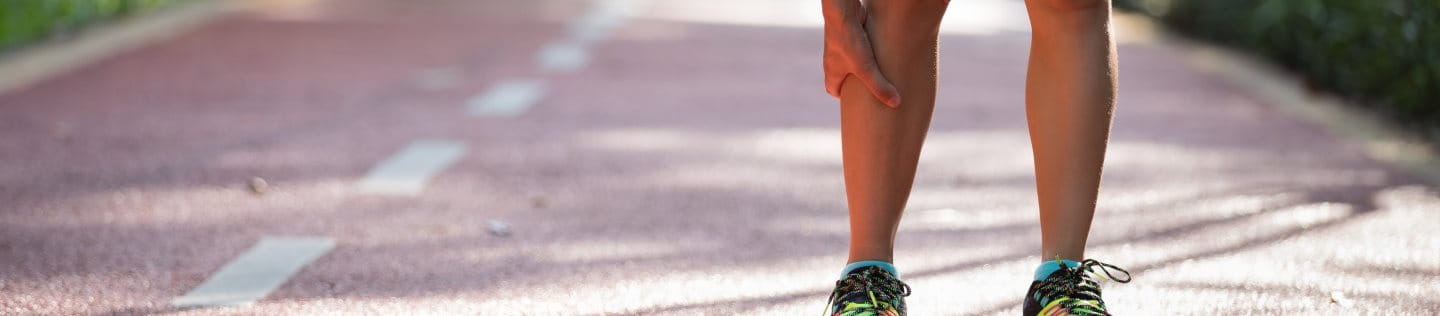 Eine Frau im Sportoutfit steht vornüber gebeugt auf einer Landstraße und hält sich ihr schmerzendes Läuferknie.