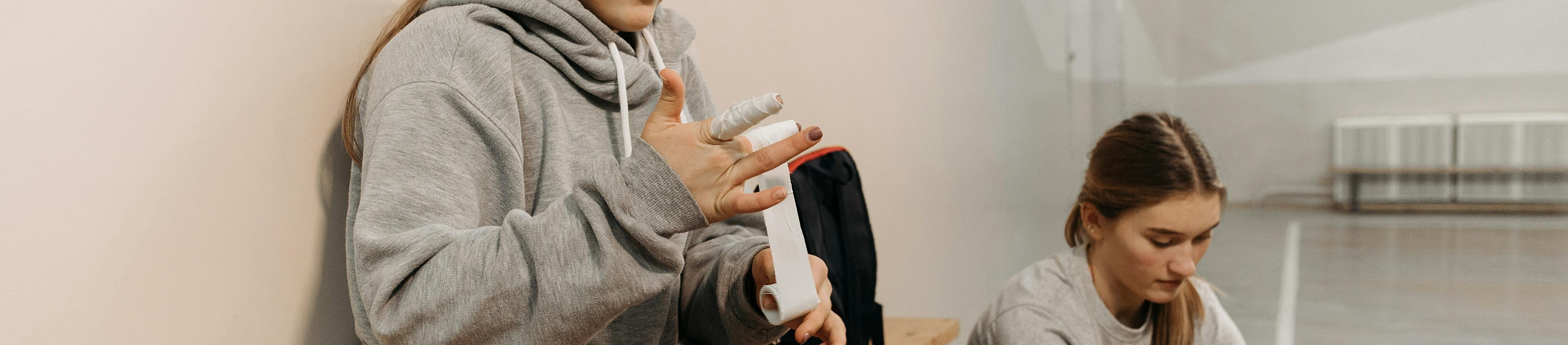 Zwei Mädchen in einer Turnhalle. Eine davon wickelt ihre Hände mit Kinesio Tape ein.